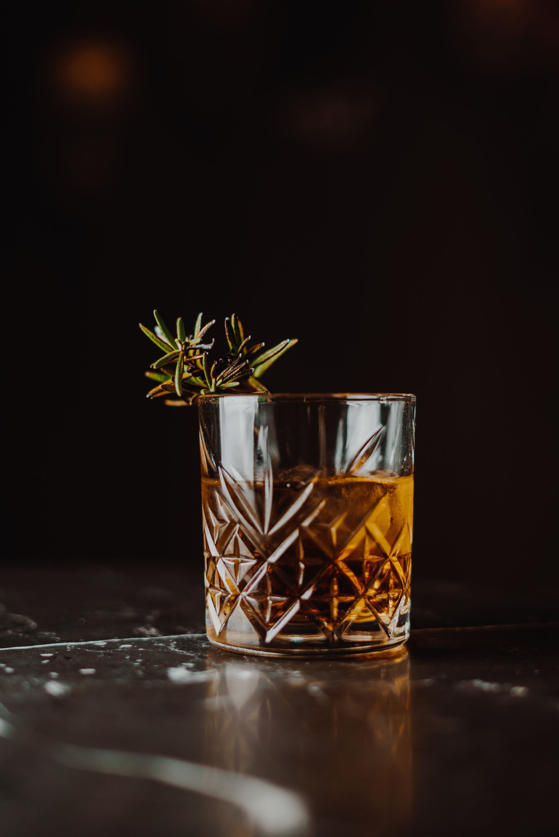 A whisky glass with rosemary on a reflective black surface